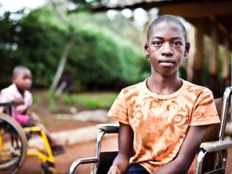 Kind im Rollstuhl schaut aufmerksam in die Kamera. Im Hintergrund sieht ein weiteres Kind im Rollstuhl