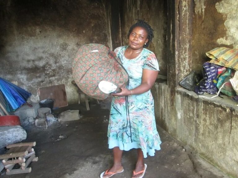 Frau präsentiert den KlimaKocher. Im Hintergrund kann man die traditionelle Kochstelle sehen. Diese befindet sich auf dem Boden und besteht aus drei Steinen und einem offenen Feuer.